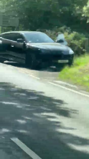 A black car parked partially off a rural road with its driver's side door open.