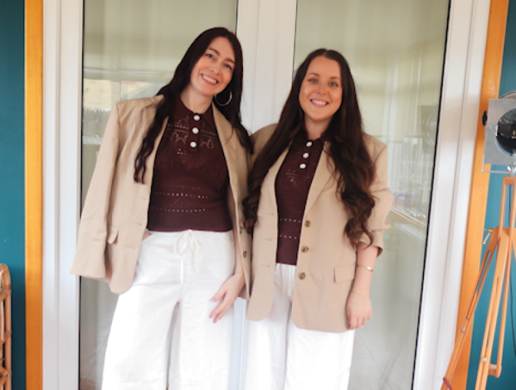 Two women wearing matching brown tops, beige blazers, and white pants, standing side by side.