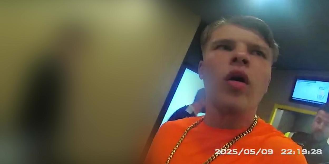 Still from a video showing Sonny Boyes in an orange shirt and gold chain, with his mouth slightly open, looking confused during his arrest.