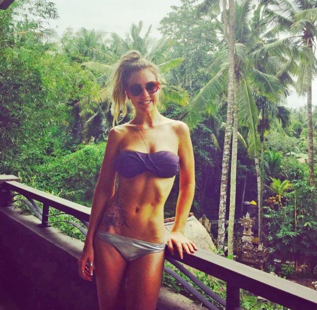 A woman in a purple bikini top and gray bottoms smiles on a balcony with a jungle in the background.