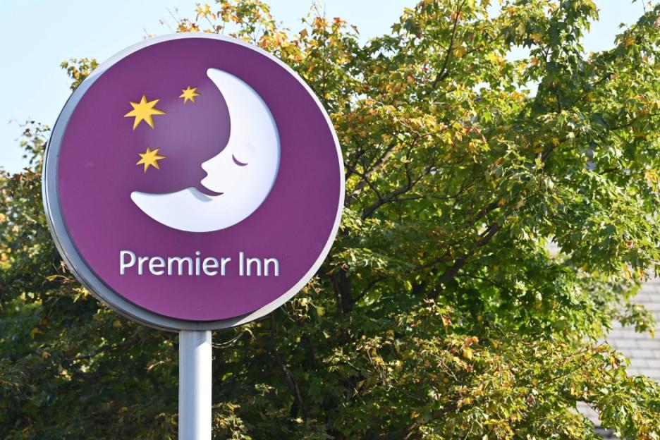 A purple Premier Inn sign with a sleeping moon and stars, against a backdrop of green trees.