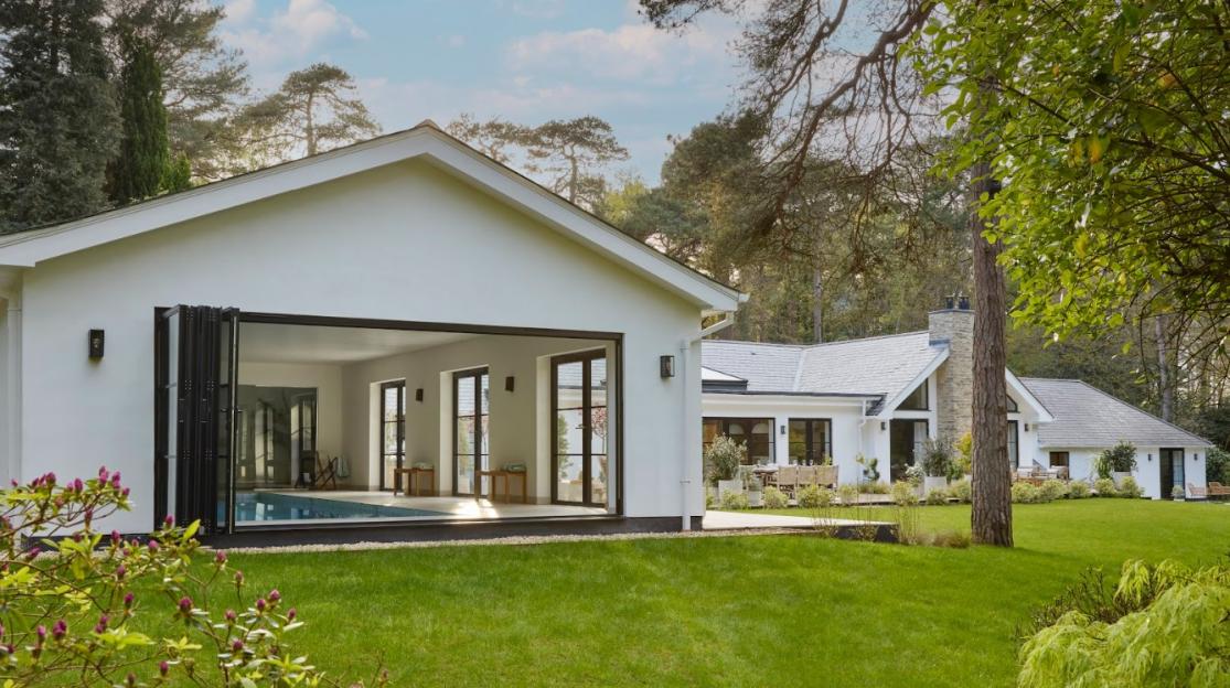 A white house with an indoor swimming pool visible through large folding glass doors, next to a large green lawn and trees.