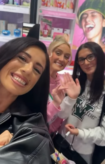 Three women in a store, one in a black leather jacket in the foreground, one in a pink top waving, and another in glasses and a white sweatshirt.