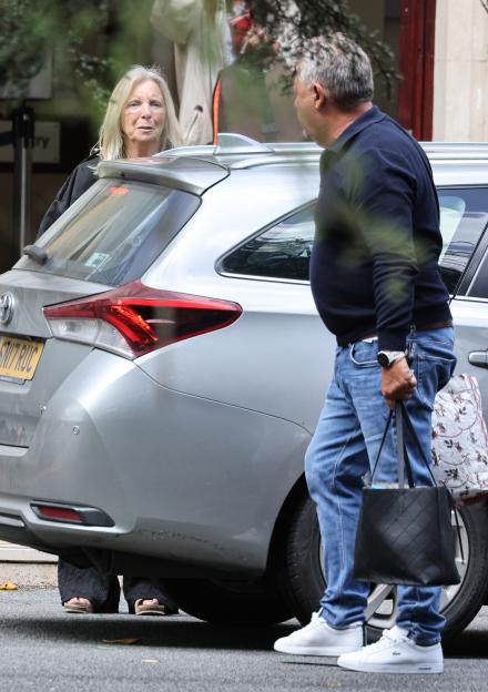 Kim Benstock, a woman with long blonde hair, standing behind a silver car, and Steve Benstock, a man in a navy polo shirt and jeans, holding a bag next to the car.