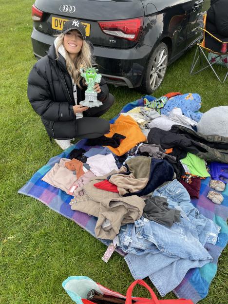 Becky Chorlton, a car boot expert, crouches on grass next to a black car, holding a green M&M's Statue of Liberty figurine, surrounded by clothes on a plaid blanket.