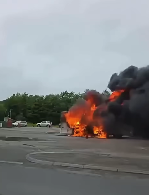 A campervan ablaze at a petrol station forecourt, emitting thick black smoke.