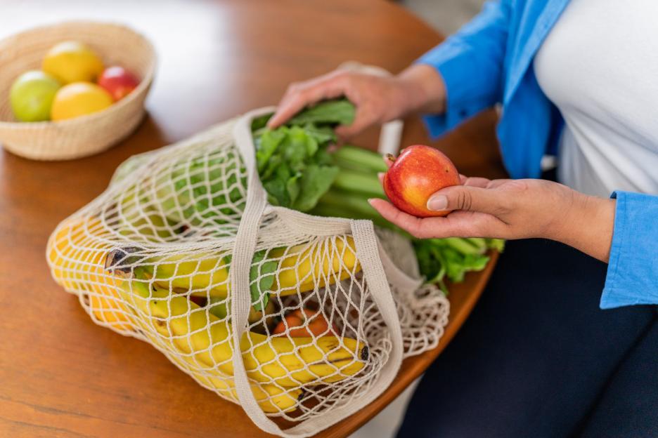 A person placing an apple into a mesh bag filled with produce.