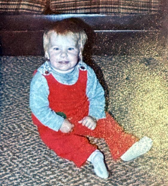 Toddler in red overalls and a light blue long-sleeved shirt sitting on a patterned carpet.