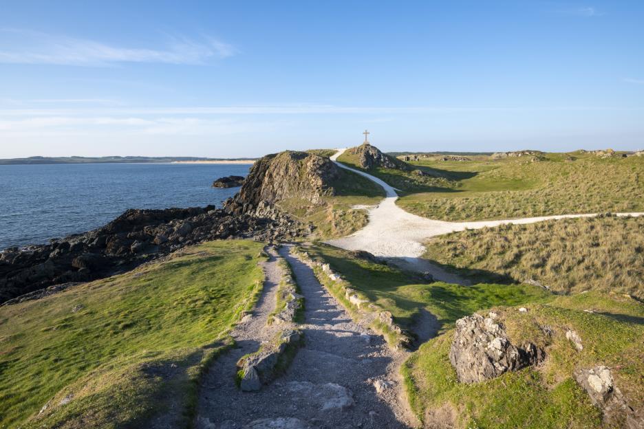 Coastal path on a sunny day in Wales, with a distant cross on a hill.