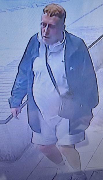 A male suspect with blonde hair, wearing a navy jacket, white polo shirt, white shorts, and a gray crossbody bag.
