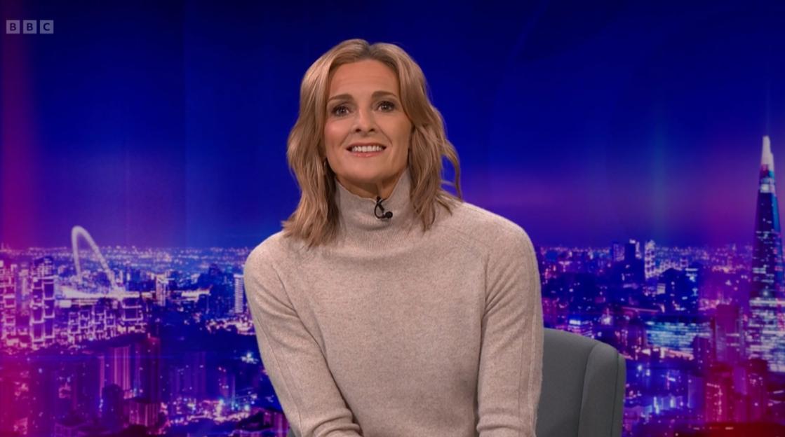 A woman in a beige turtleneck sits in front of a blue and purple city skyline backdrop.