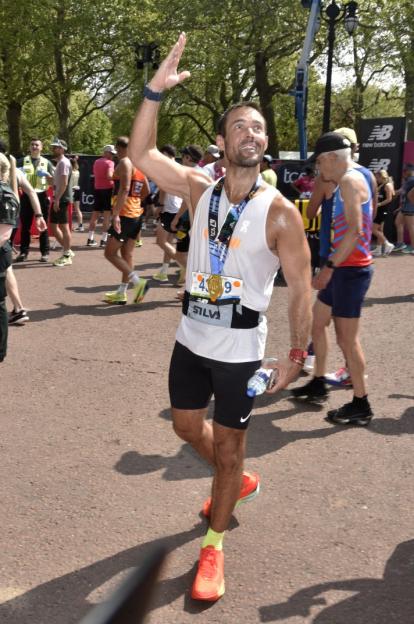 A man, wearing a white singlet and black shorts, waves while holding a medal after completing the London Marathon.