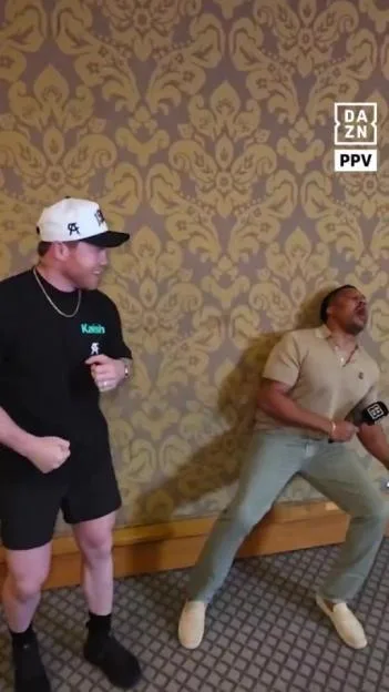 A man wearing a black t-shirt and white hat pretends to punch another man who leans back against a patterned wall, holding a microphone.