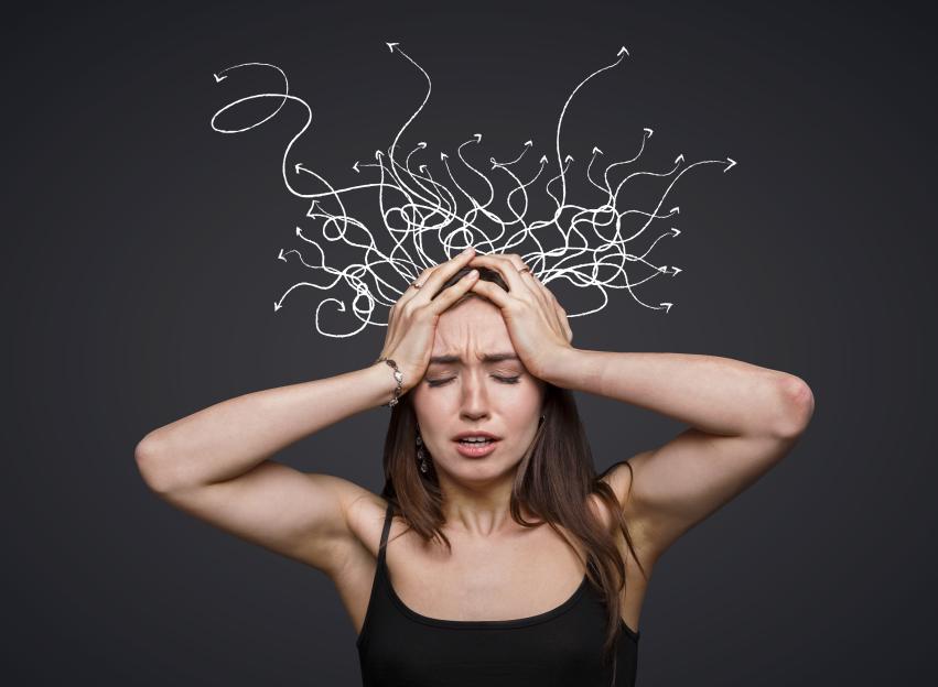 Composite image of a woman holding her head with tangled white lines and arrows symbolizing overwhelming thoughts emerging from her head against a gray background.