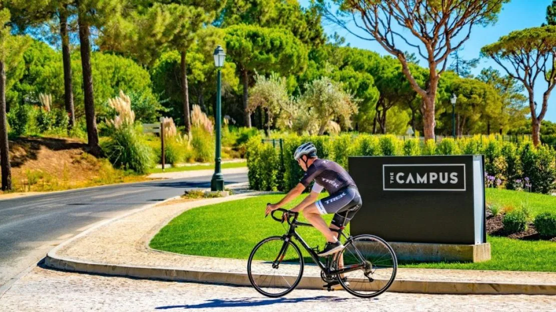 A man in cycling gear rides a bicycle on a paved path next to a sign for "THE CAMPUS" in Quinta do Lago.