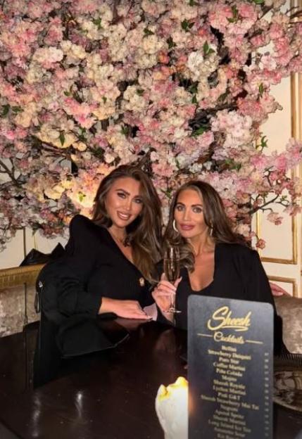 Two women pose for a photo in front of a wall of pink flowers, one holding a glass of champagne.