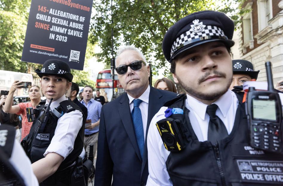 Huw Edwards, former BBC presenter, arrives at Westminster Magistrates' Court, accompanied by police officers, with a protest sign visible in the background.
