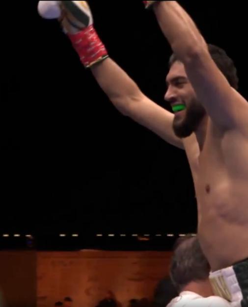 A shirtless boxer with a beard and a green mouthpiece smiles with his arms raised in victory.