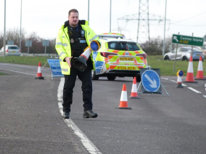 A police officer in a high-visibility jacket holds a traffic cone, with a police vehicle and additional traffic cones visible in the background, indicating an emergency scene.