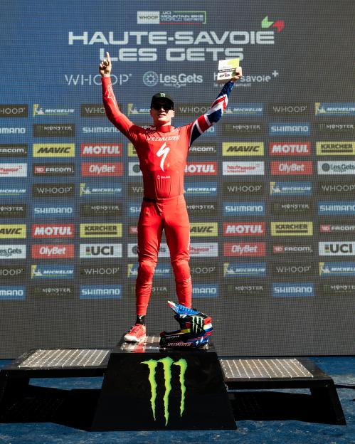 A mountain biker in red race gear, standing on the top podium step, raising their arms in victory at the UCI Mountainbike World Series.