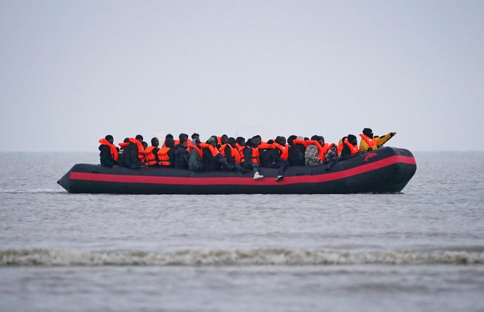 Migrants in a small inflatable boat with orange life jackets in the water.