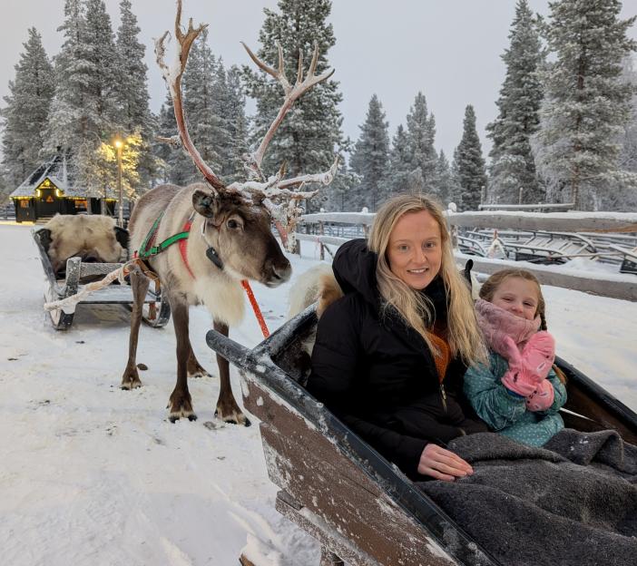 A woman and a child sitting in a wooden sleigh pulled by a reindeer in a snowy forest.