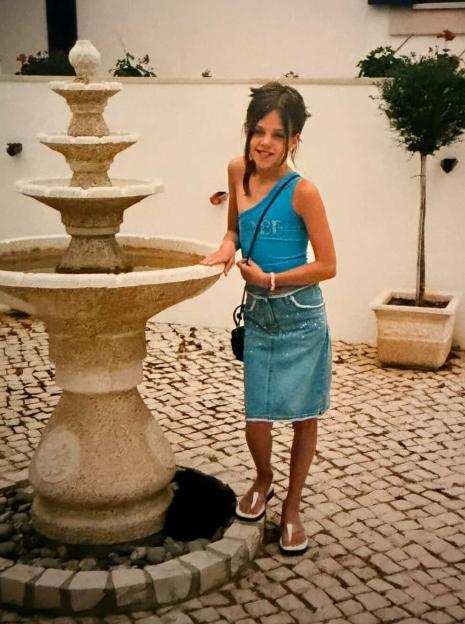 A young girl in a blue top and denim skirt stands next to a stone fountain.