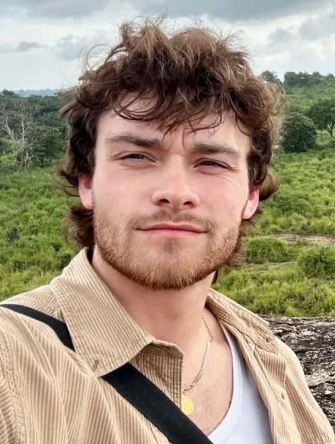 Close-up of a young man with curly brown hair and a beard, wearing a tan corduroy shirt and a gold chain, with a green, hilly landscape in the background.