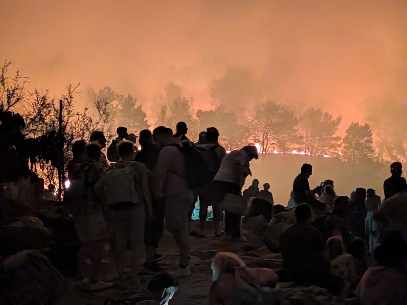 Silhouettes of people stand in the foreground as a wildfire burns in the forest behind them under a smoke-filled, orange sky.