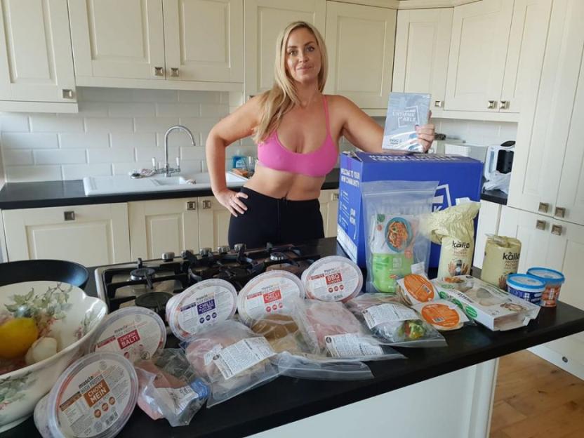 A woman in a pink sports bra holds up a book next to a counter full of meal prep items.