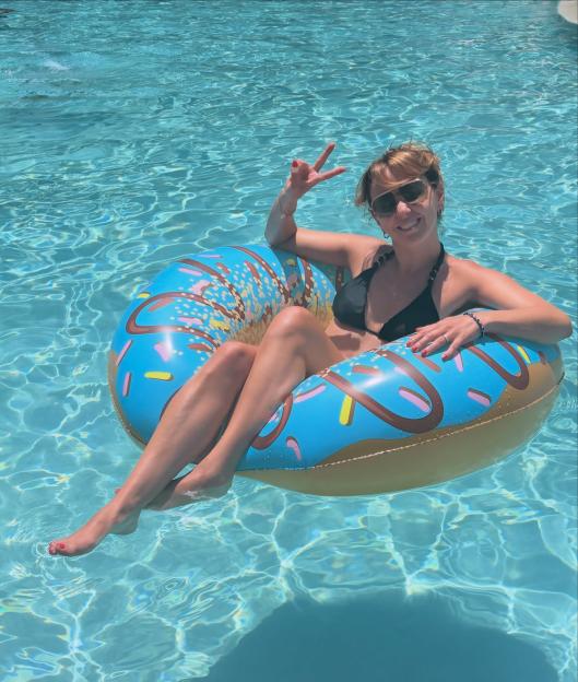 Samia Longchambon in a black bikini, sitting in a donut-shaped inflatable float in a swimming pool, making a peace sign with her hand.