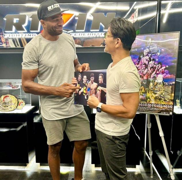Two men, one in a RIZIN hat, holding a photo of himself with other fighters, speaking in front of a RIZIN banner and event poster.