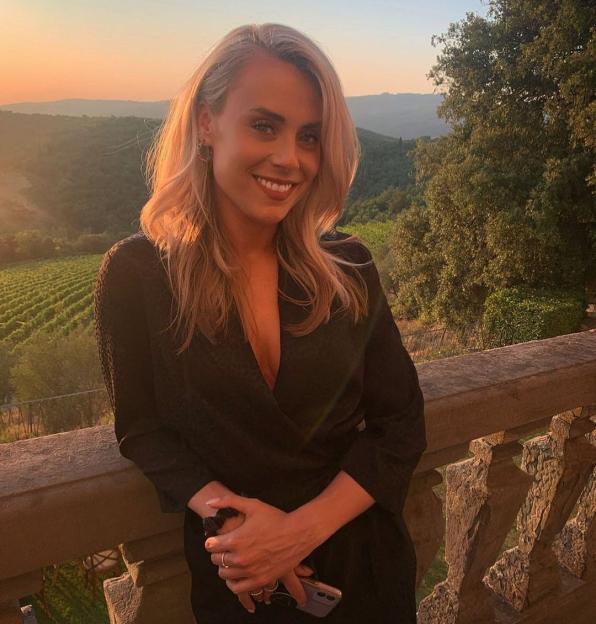 A smiling woman in a black dress leans on a stone balcony railing, with a vineyard and sunset sky in the background.