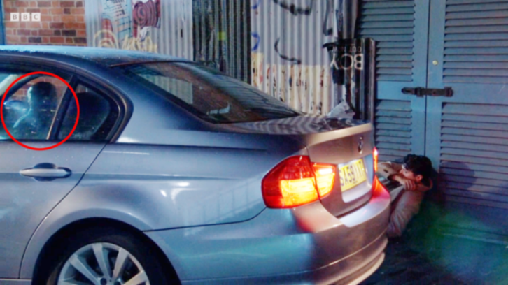 A man with dark hair in a light brown coat huddles on the ground behind the rear of a silver car as a reflection of a crew member is visible in the car's window.