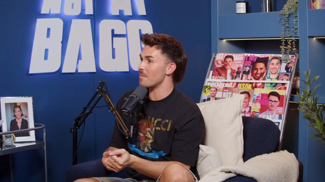A man seated at a podcast with magazines featuring his face in the background.