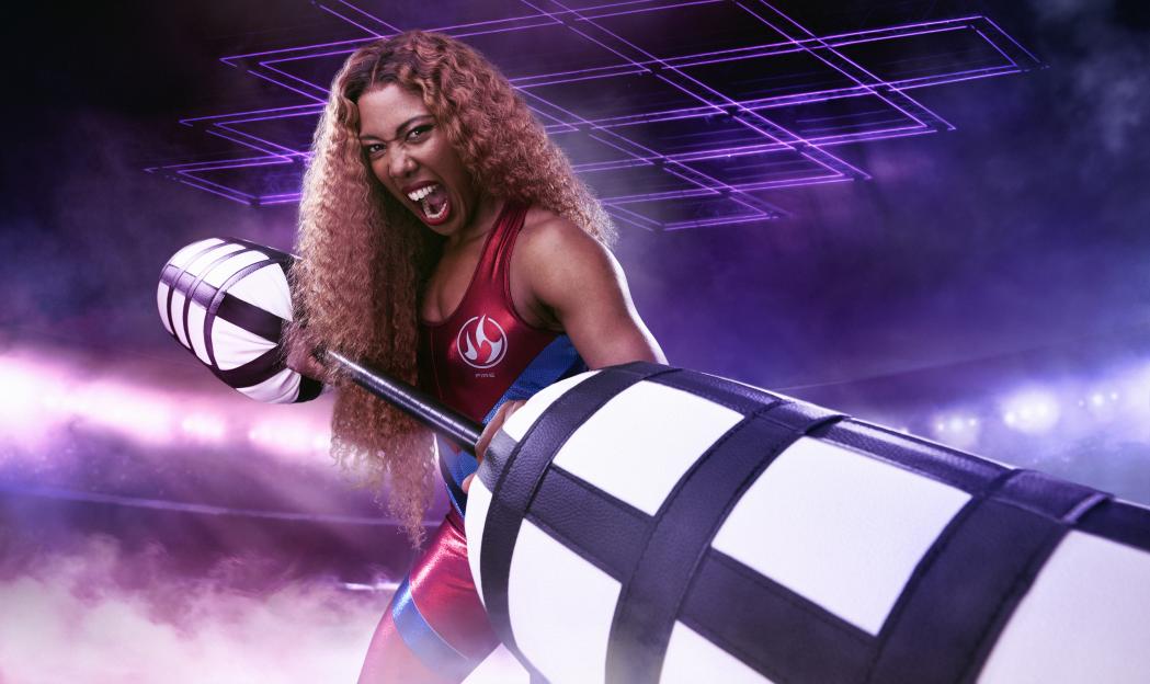 Fire from Gladiators, a woman with curly brown hair, shouts while holding two black and white padded jousting sticks.