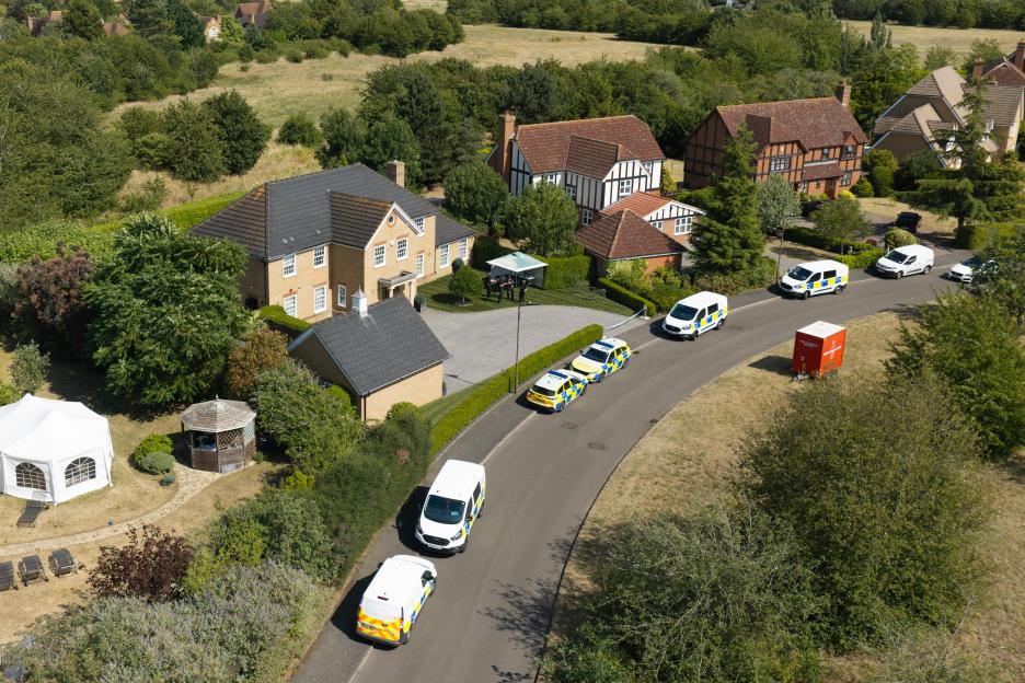 Aerial view of a residential street lined with houses, trees, and several police vehicles, with a white tent in front of one house.