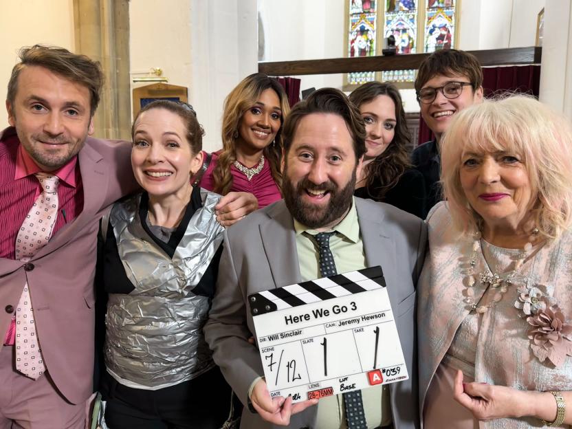 Cast members of "Here We Go S3", including Tom Basden, Katherine Parkinson, Tori Allen-Martin, Jim Howick, Freya Parks, Jude Collie, and Alison Steadman, pose for a group photo with a film clapperboard.