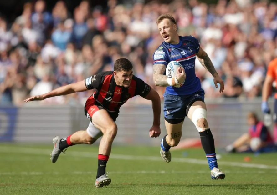 Tom Johnstone running with the ball during a rugby match with a defender in pursuit.