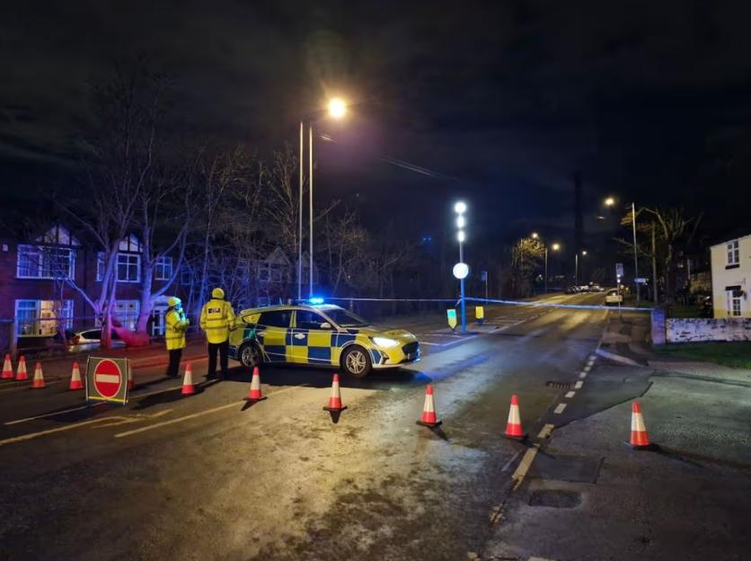 Police car with flashing blue lights and two officers in yellow vests block a road at night with orange cones and blue tape.