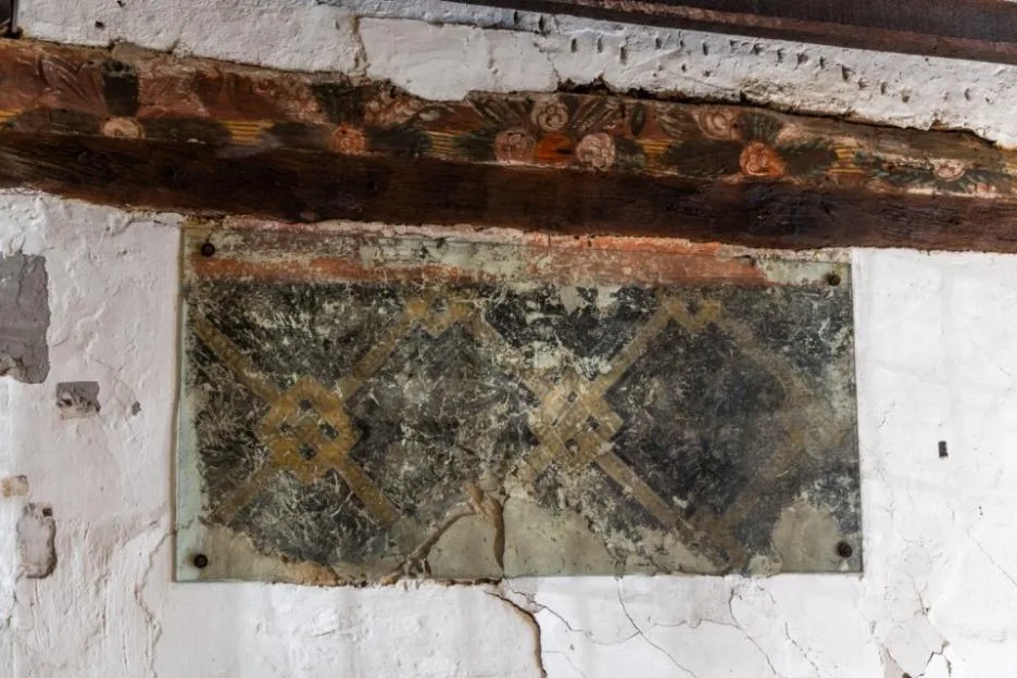 Faded medieval wall paintings protected by a glass pane under a wooden beam.