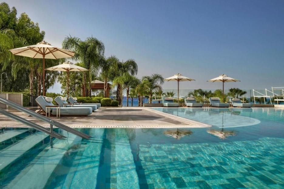 A resort swimming pool with lounge chairs and umbrellas, palm trees, and the sea in the background.