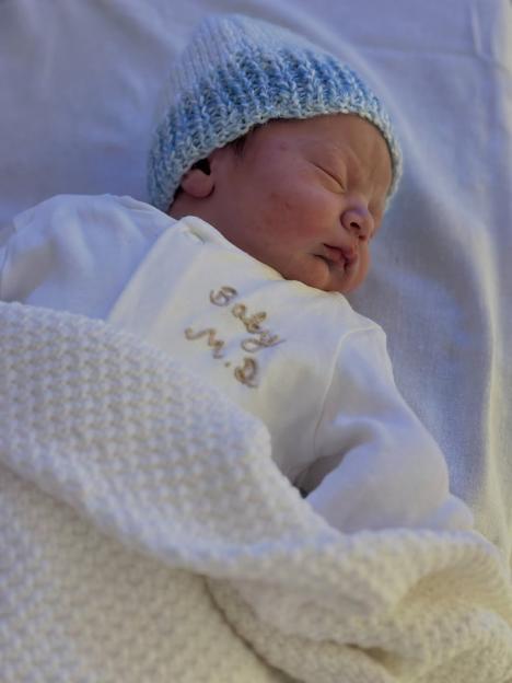 Newborn baby sleeping under a white blanket, wearing a blue and white knit hat and a white shirt with "Baby M.S" embroidered on it.