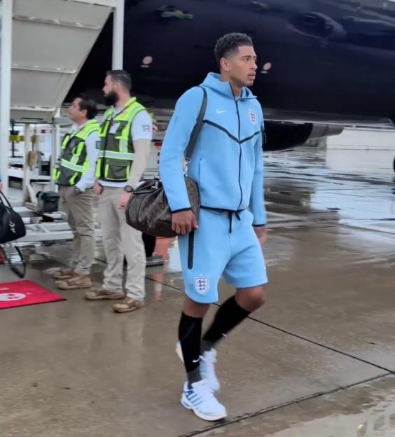 Jude Bellingham wearing a light blue Nike tracksuit with an England crest, carrying a duffel bag, walking away from an airplane with airport staff in the background.