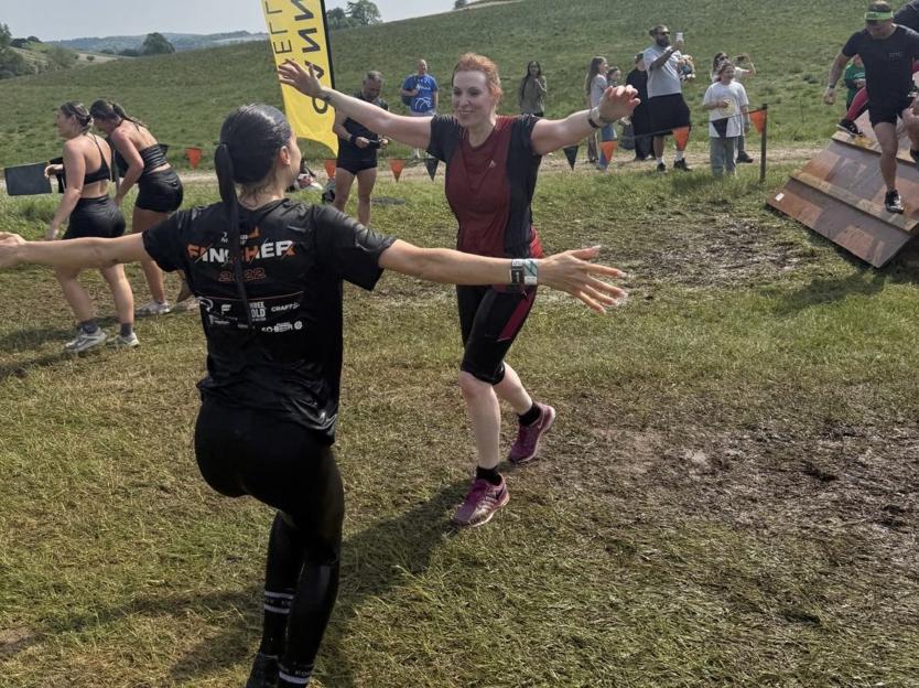 Two women, one in a red and black shirt and one in a black "Finisher 2022" shirt, approach each other with arms outstretched on a muddy, grassy course.