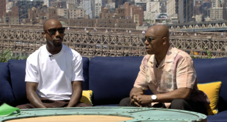 Two men in sunglasses sitting on an outdoor couch with a city skyline in the background.