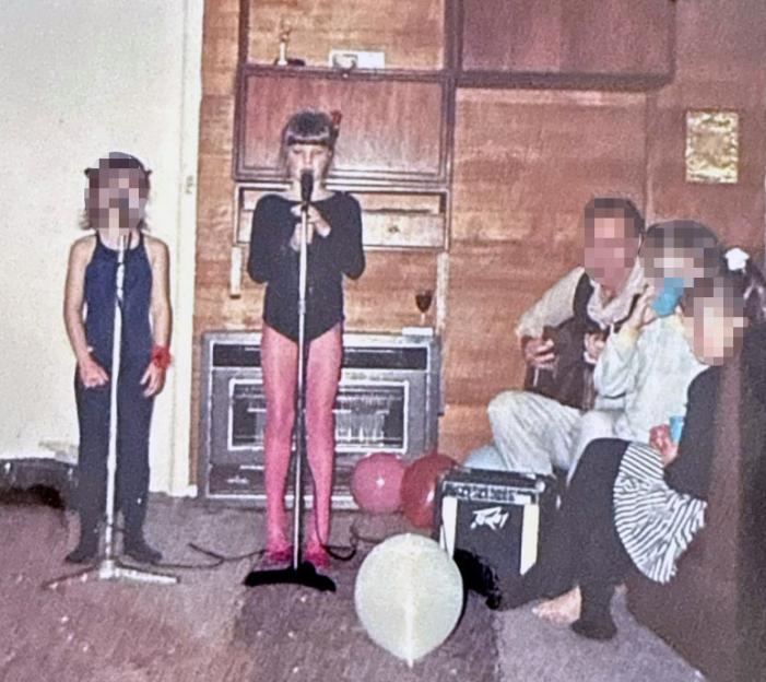 Two blurred young girls singing into microphones while a blurred man plays guitar and two blurred children watch.