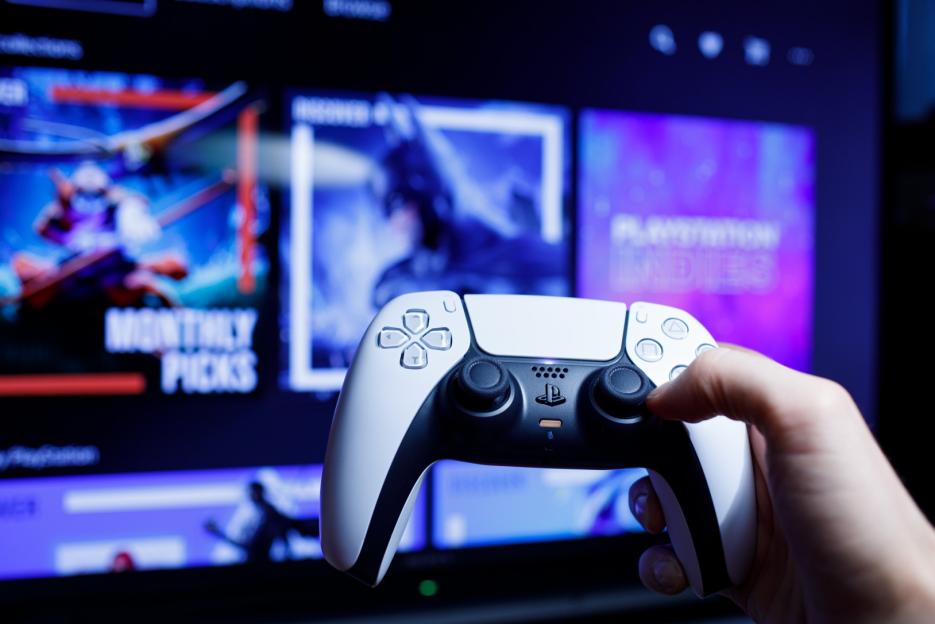 A hand holding a white and black PlayStation 5 controller in front of a TV displaying game titles.