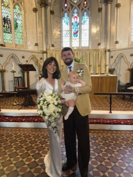 A family in a church with the father holding a baby, and the mother holding a bouquet of white flowers.