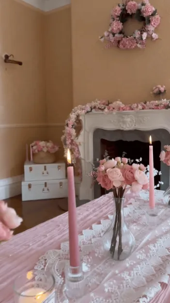 A dining table decorated with pink candles, a vase of pink flowers, and a white lace runner, set against a backdrop of a fireplace adorned with pink floral garlands and a matching floral wreath on the wall.